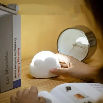 White cloud-shaped night light with soft glow, held in hand beside books and lamp