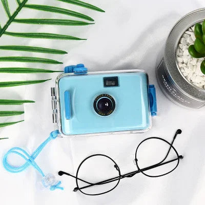 Light blue instant camera with blue strap, lens, and buttons, styled with glasses and greenery
